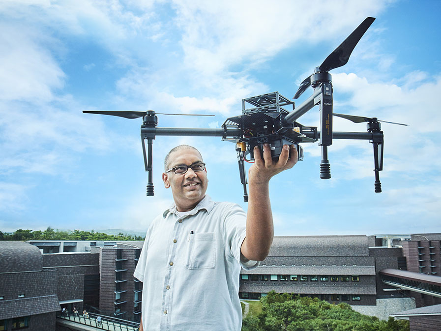 An indian man stands on the roof of a laboratory with the blue sky behind him, holding a drone.